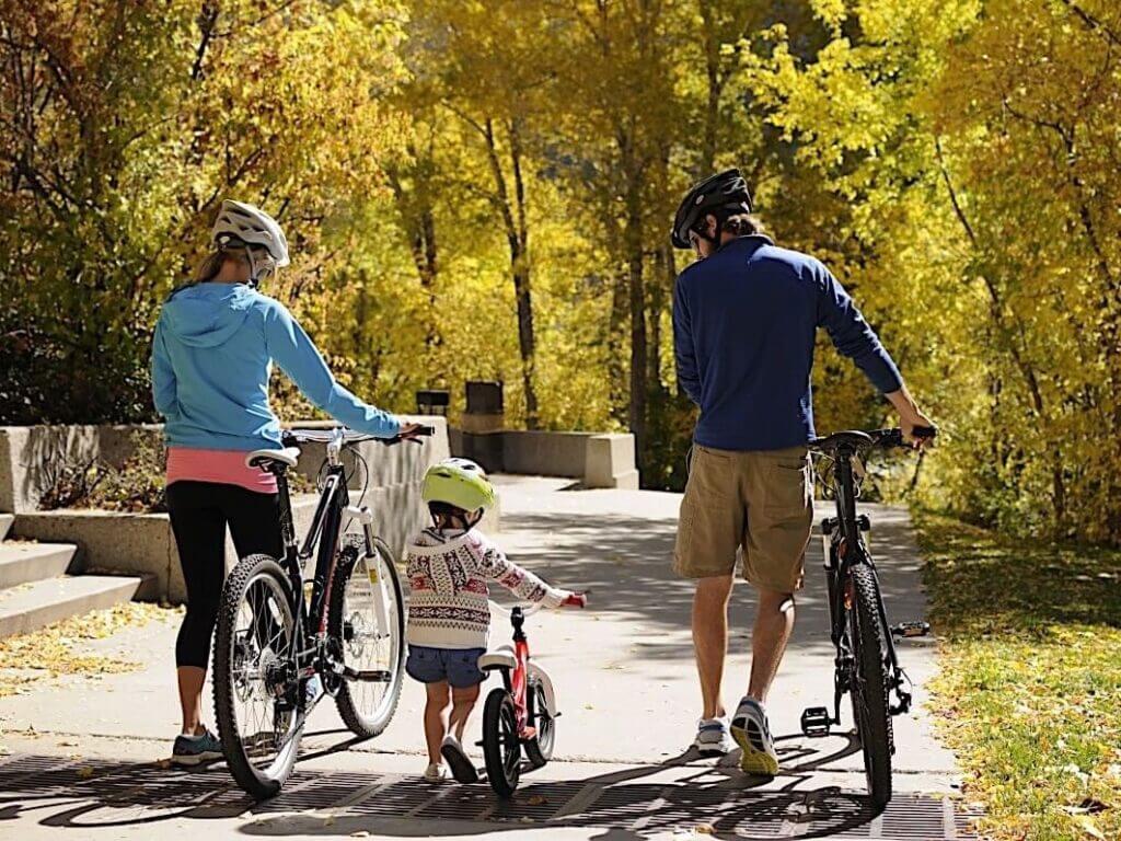 A family of three, wearing helmets, walk their bikes along a sunlit path in a park surrounded by the vibrant fall colors of golden autumn leaves. The young child, in the center, rides a small bicycle.