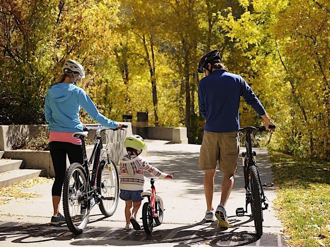 A family of three, wearing helmets, walk their bikes along a sunlit path in a park surrounded by the vibrant fall colors of golden autumn leaves. The young child, in the center, rides a small bicycle.