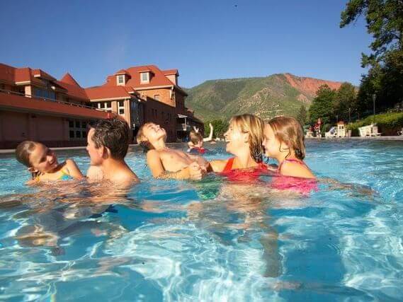 A family of five is delighting in a sunny spring break day at a large outdoor pool. Two adults and three children are playing and smiling at each other, while the backdrop boasts buildings and a mountain landscape under a clear blue sky.