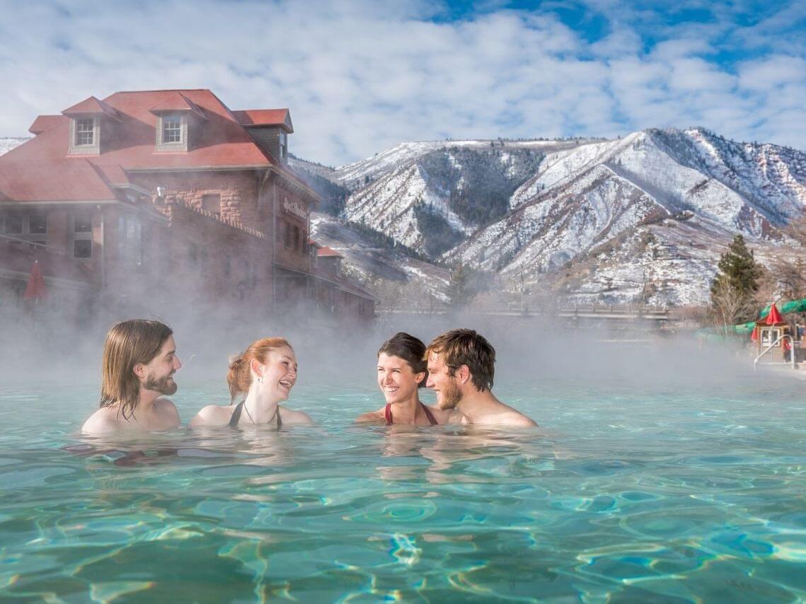 Four people enjoy an outdoor hot spring pool, surrounded by mist. In the background, there's a rustic building and snow-covered mountains under a partly cloudy blue sky.