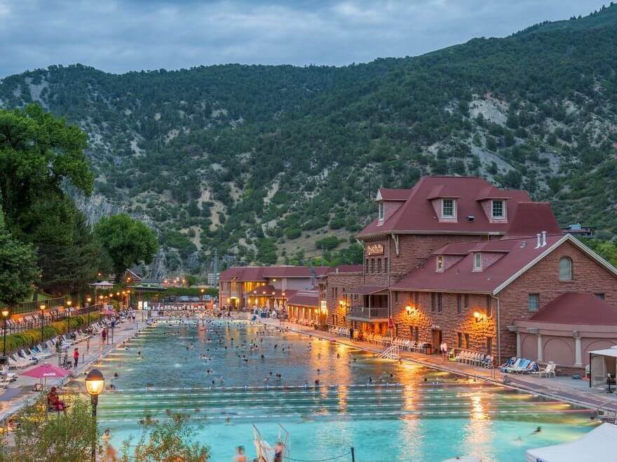 A large outdoor pool buzzes with activities as people swim and lounge, set against a mountainous backdrop. Warm lights illuminate the area and a historic brick building. The sky is cloudy, creating a serene atmosphere.