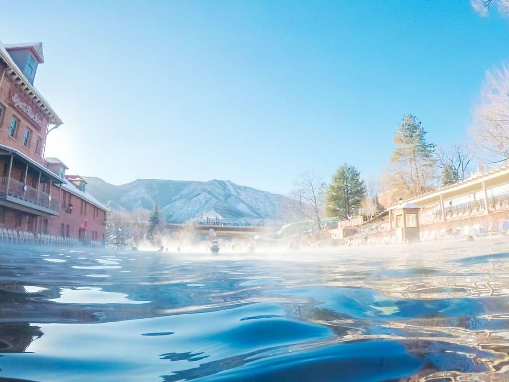 A view from the water reveals a large outdoor pool surrounded by historic buildings and trees. Steam rises as you soak away your worries, while snow-capped mountains stand majestically in the background under a clear blue sky.