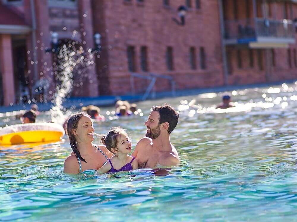 A family of three enjoys swimming in a large outdoor pool, where the mother and father are holding their smiling young daughter. The pool area is bustling with other swimmers making merry splashes, while a fountain sprays water gleefully in the background.