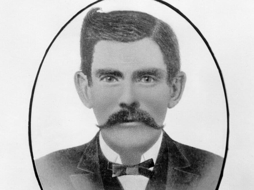 Black and white portrait of a man with a thick mustache resembling Doc Holliday, wearing a dark suit, white shirt, and bow tie. The image is framed in an oval shape.