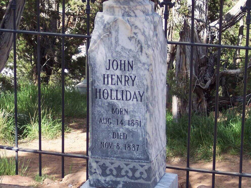 A marble tombstone engraved with the name John Henry Holliday, famously known as Doc Holliday, born August 14, 1851, died November 8, 1887. It is set against an iron fence with trees and grass in the background.