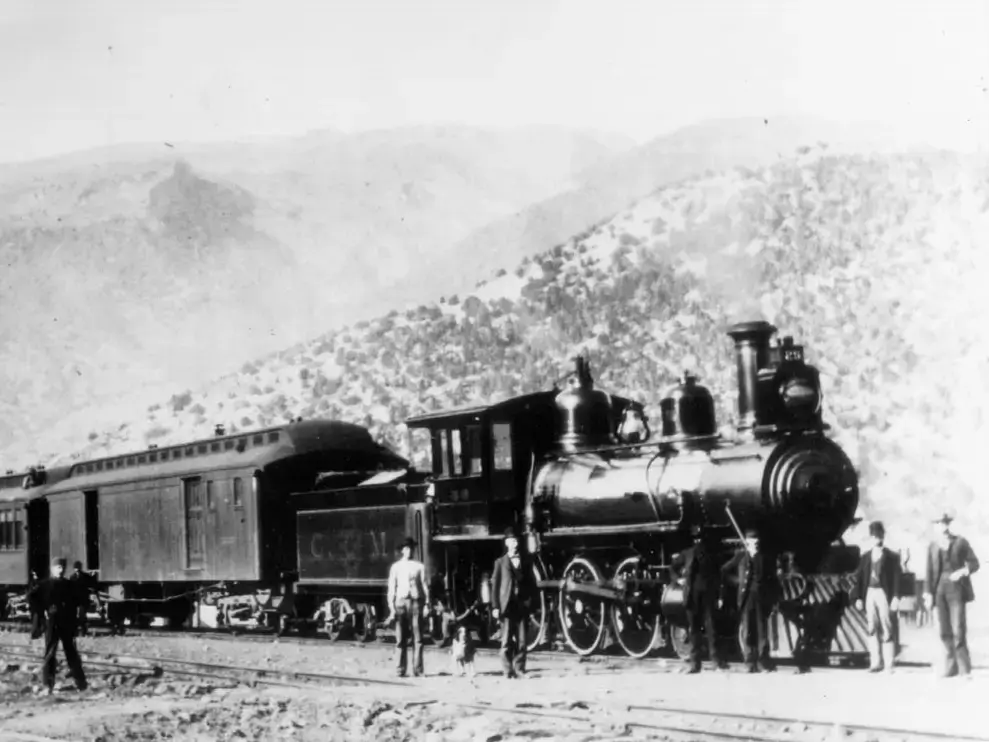 Black and white photo of an old steam locomotive with several men posing around it, situated on a track in the mountainous terrain near Union Station. One of the train cars bears the marking C.P.R.R., signifying its connection to the Central Pacific Railroad.