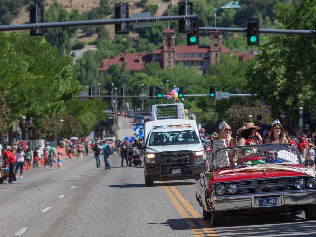 A parade celebrating Strawberry Days features people in costumes riding a red convertible car, followed by a decorated white truck. Spectators line the street under a sunny sky, with trees and buildings providing a vibrant backdrop.