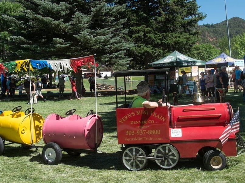 A small red and yellow train with Verns Mini Train written on it chugs along the grass on a sunny Strawberry Days festival. The train, adorned with colorful flags and an American flag, passes by vendor booths and happy people under tall trees in the vibrant background.