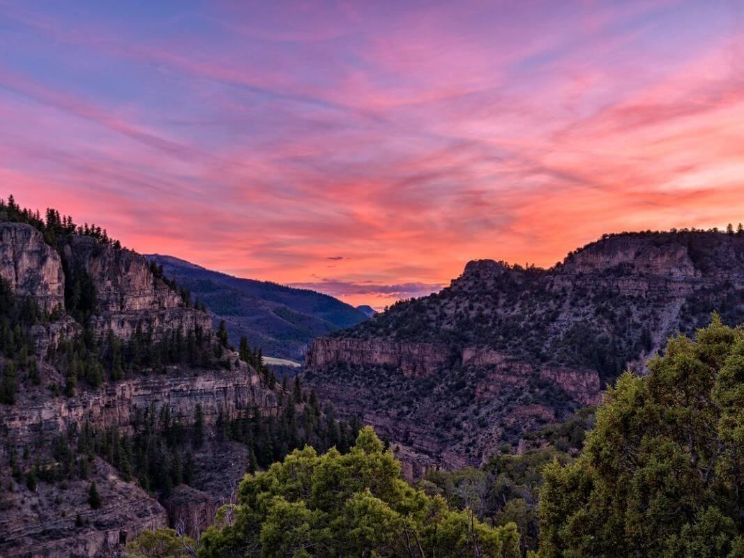 A breathtaking sunset over a rugged canyon landscape, where silhouetted trees hint at the freshness of spring. The mountainous terrain complements a sky painted in vibrant hues of pink, orange, and purple, creating a stunning contrast against the darkened land below.