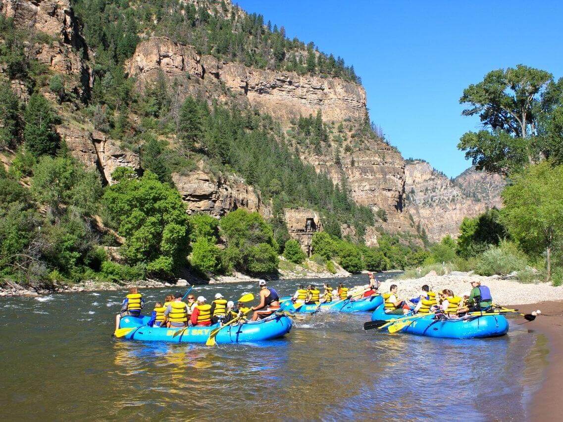 A group of people in life jackets are rafting in blue rafts on a river, embraced by lush green trees and towering rocky cliffs under the clear summer sky. The scene conveys a sense of adventure and nature.
