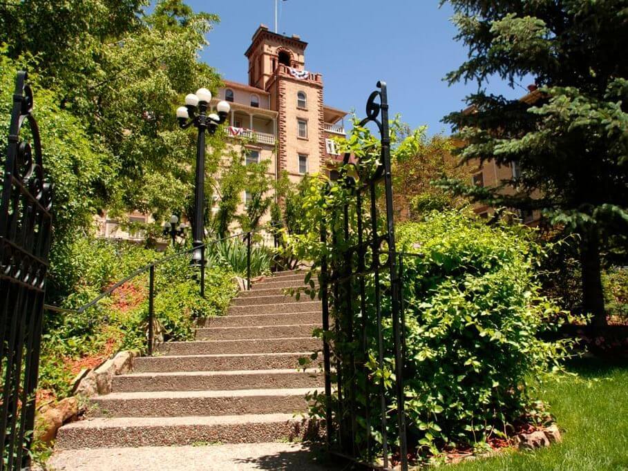 A stone staircase surrounded by lush green plants leads up to a historic building with a tower and balconies, offering plenty of activities for visitors. Black wrought iron lampposts and an ornate gate frame the pathway, all under a clear blue sky.