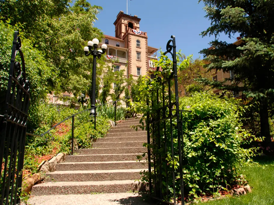 A stone staircase leads up to a historic brick building with a clock tower, offering perfect photo opportunities. The scene is surrounded by lush green trees and shrubs under a clear blue sky. A wrought iron gate elegantly frames the entrance to the pathway.