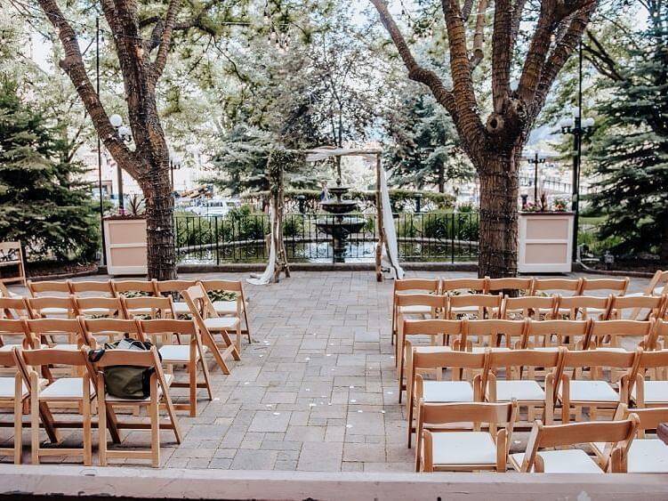 An outdoor wedding ceremony setup, meticulously planned, features rows of wooden chairs facing an altar adorned with white fabric, positioned between two large trees. A lush garden background with a decorative fountain completes the romantic ambiance.