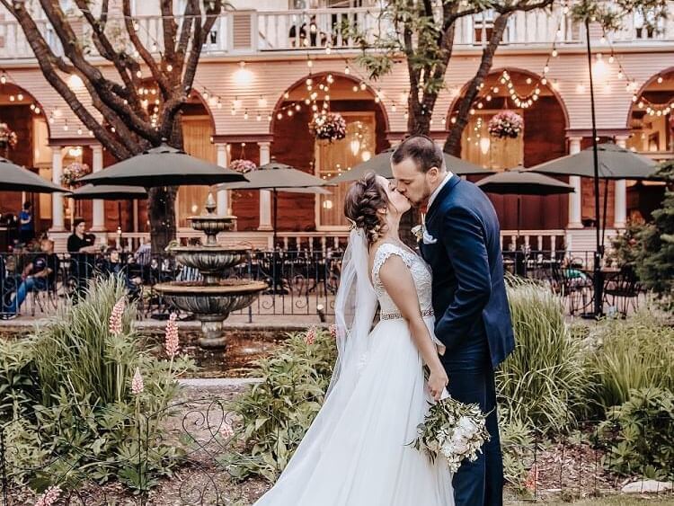 In a beautifully planned garden courtyard, the bride and groom share a kiss. The bride, in her white gown, holds a bouquet as the groom complements her in his navy suit. String lights and lush greenery envelop them, crafting an enchantingly romantic atmosphere.