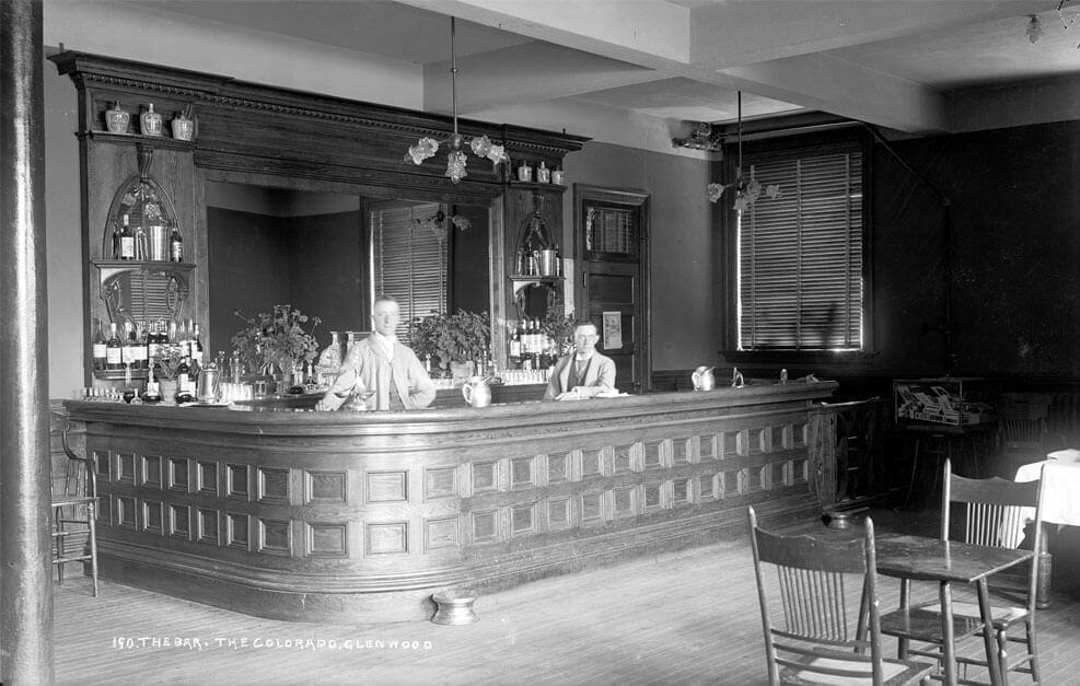 A vintage black and white photo captures two bartenders behind a large, ornate wooden bar at Hotel Colorado