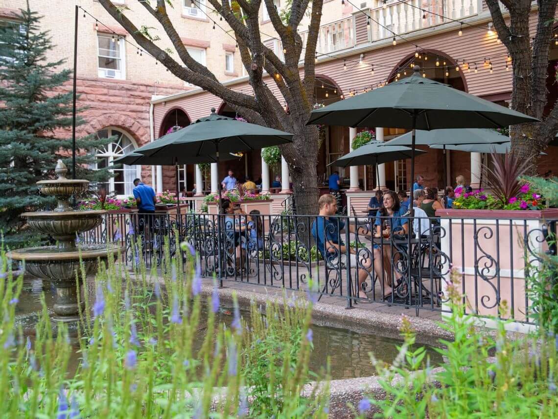 People sit at outdoor tables under umbrellas in a charming courtyard with a fountain, a locals' choice for relaxation. Surrounded by greenery and string lights, the setting exudes a relaxed atmosphere. The backdrop features a historic brick building with arched windows.