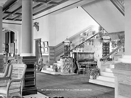 Black and white image of a spacious, stylish interior featuring a grand wooden staircase linked to a vintage piano and decorative plants. The area is adorned with framed art and intricate architectural details. The text reads The Grand Stairway, The Glenwood.