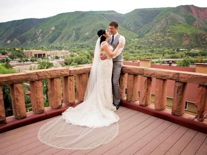 An enchanting wedding unfolds as the bride in a white dress and veil shares an intimate moment with the groom in a suit and tie on a wooden balcony, framed by lush green mountains and a charming townscape.