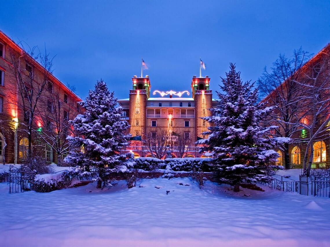 A historic hotel, elegantly adorned with Christmas lights, glows warmly against a snowy evening backdrop. Snow-covered trees and bushes line the path leading to the entrance, creating a festive and serene winter scene filled with holiday spirit.