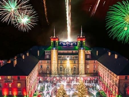 A grand building is illuminated at night with colorful lights, embracing cherished holiday traditions. Fireworks burst in the sky, displaying vibrant colors. Trees in front are adorned with sparkling lights, enhancing the festive atmosphere.
