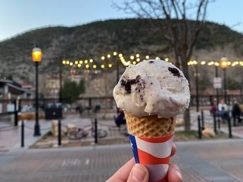 A hand holds a cookies and cream ice cream cone, its patriotic stars and stripes contrasting the warm hues of a park at dusk. String lights twinkle gently above, with a distant hill completing the scene—a perfect escape from the heat of the day.