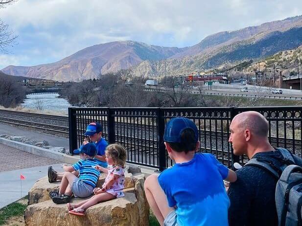 A group of people, including three children and two adults, sit on rocks near a fence by the railroad tracks. It's spring break, and in the background, a river flows with mountainous terrain beneath a cloudy sky. They seem to be enjoying this scenic outdoor setting.
