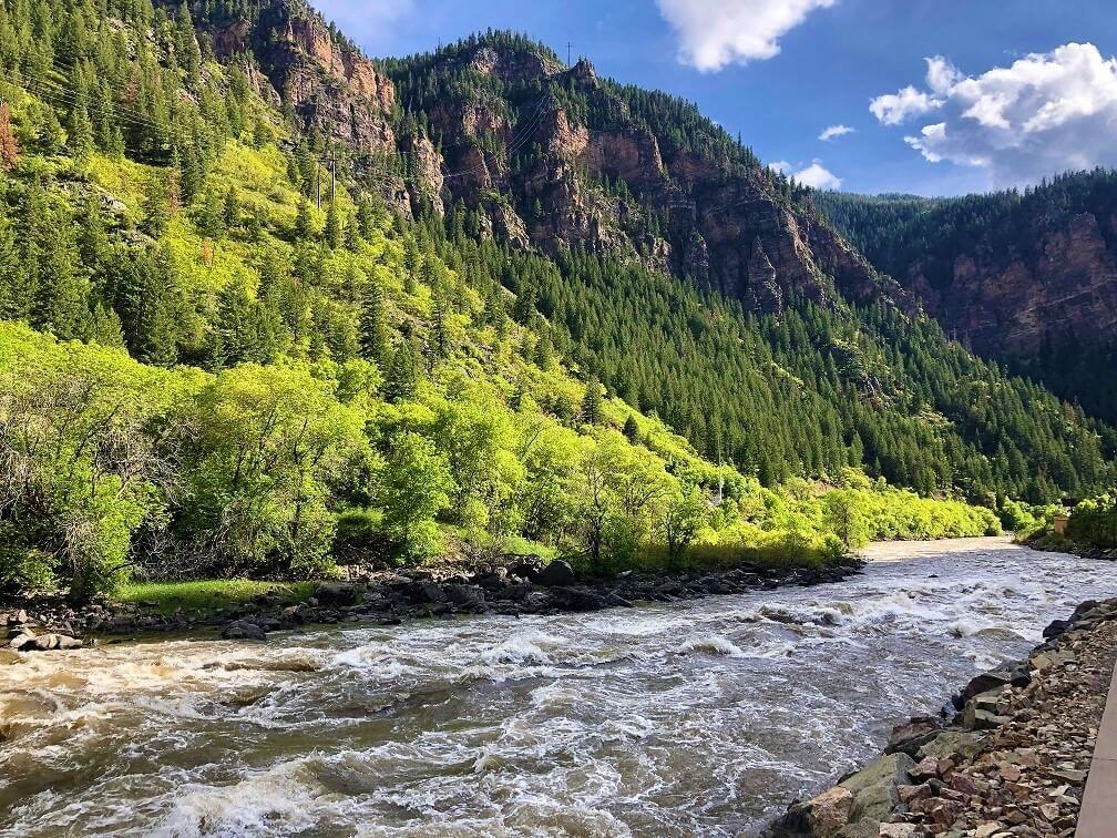 A scenic view of a river flowing swiftly through a lush, green forested canyon with rocky cliffs. The sky is blue with scattered clouds, sunlight highlighting the vibrant greenery and rugged terrain—perfect for fly fishing amidst nature’s splendor.