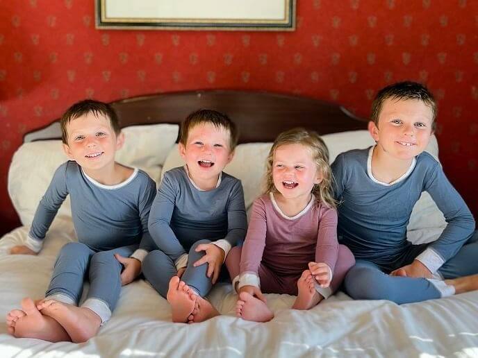 Four children are sitting on a bed, smiling during spring break. Three wear blue pajamas, while one child wears pink. The room features a red patterned wall and a wooden headboard. They appear happy and relaxed.