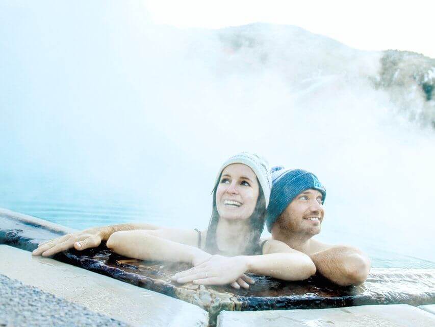 A couple in winter hats relaxes, soaking away their worries in a steaming hot spring pool. They lean on the edge, smiling and gazing into the distance as mist rises around them, with snowy mountains providing a breathtaking backdrop.