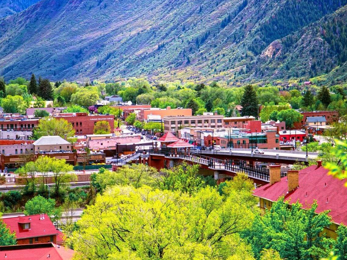 A vibrant mountain town nestled in lush greenery, featuring red-roofed buildings and a bustling main street. The towering mountains ripple with patches of greenery under a bright blue sky, creating a picturesque backdrop.
