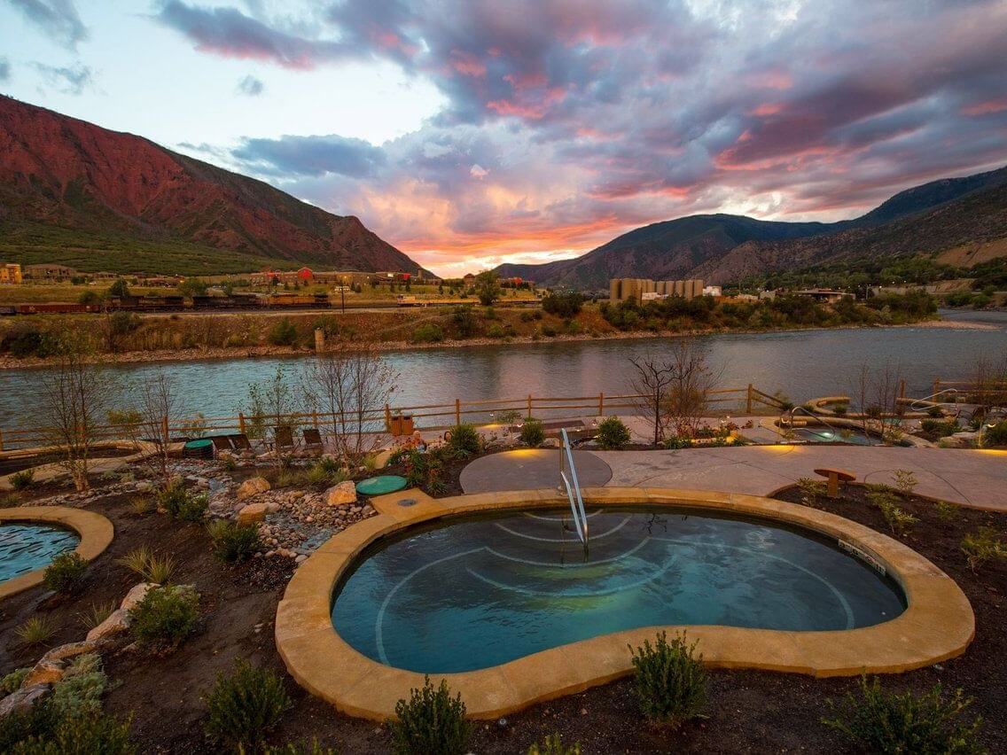 A serene outdoor hot spring pool surrounded by lush greenery offers perfect photo opportunities as it overlooks a tranquil river and mountainous landscape at sunset. The sky is painted with vibrant hues of orange, pink, and purple, reflecting beautifully on the water's surface.