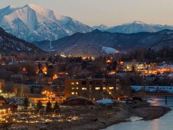 A serene evening view of a snow-capped mountain range with illuminated buildings and houses in the valley captures the essence of outdoor tranquility. The landscape features a winding river in the foreground, under a clear sky transitioning into dusk.