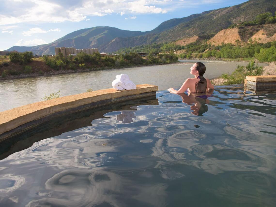 A person relaxes in an outdoor infinity pool, where gentle ripples grace the surface, overlooking a scenic river and mountains under a partly cloudy sky. A stack of white towels is placed on the edge of the pool.