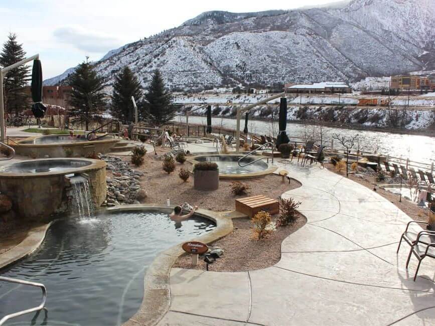 Iron Mountain hot springs resort boasts several pools and a stunning waterfall feature, set against the backdrop of snow-dusted peaks and a flowing river. Outdoor seating and umbrellas line the curved paved pathway, offering guests a serene escape.