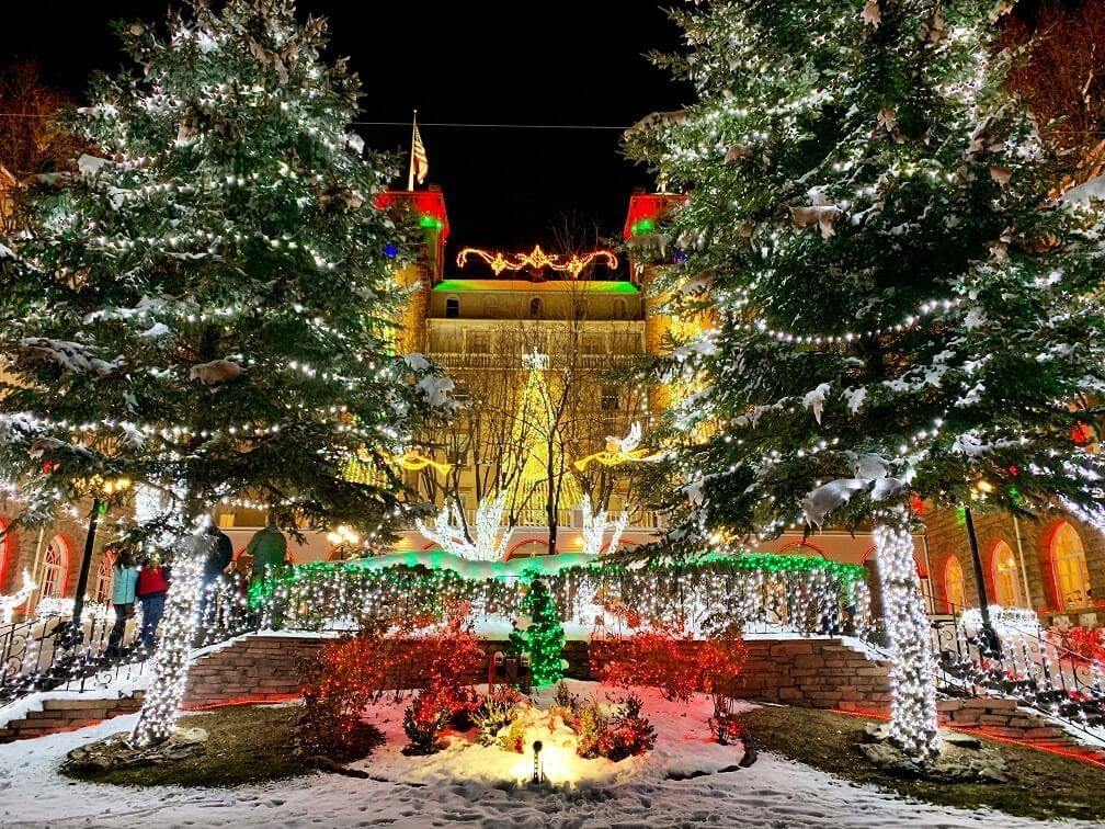 Illuminated holiday decorations reimagine the night as they cover trees and a building. Snow blankets the ground, while colorful lights create a festive atmosphere with a glowing Christmas tree centerpiece.