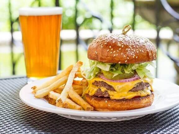 A cheeseburger with lettuce, onions, and melted cheese on a sesame seed bun sits next to a side of fries, creating perfect photo opportunities. In the background, a glass of amber-colored beer rests on a black mesh table, completing the picturesque scene.