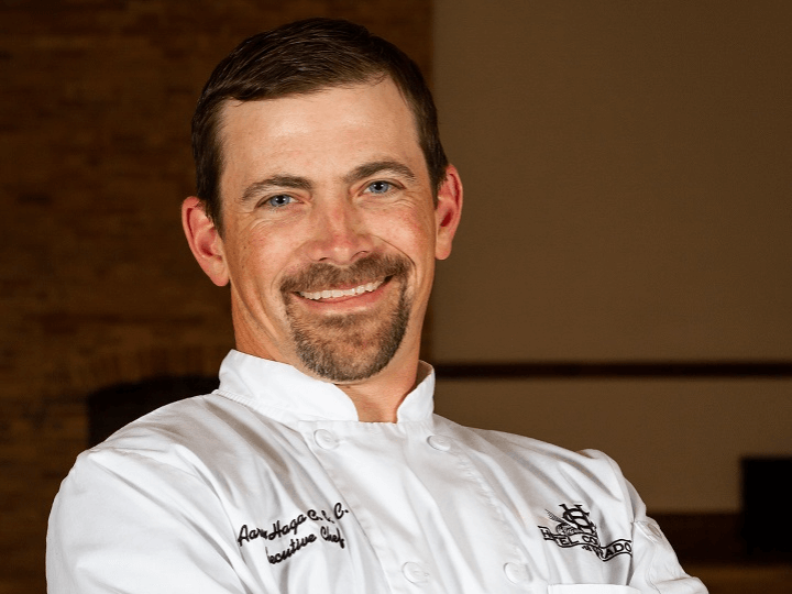 A man in a white chef's coat featuring a logo stands smiling, embodying the spirit of Thanksgiving. He has short brown hair, a goatee, and wears a nametag on his coat. The softly blurred background exudes warm tones, reminiscent of holiday gatherings.