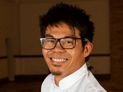 A smiling chef with spiky black hair and glasses is wearing a white chef's jacket. The background is a blurred indoor setting.