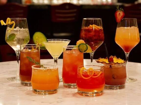 A variety of colorful cocktails displayed on a marble table in a charming courtyard. The drinks are in different types of glasses, some garnished with fruit slices, herbs, or umbrellas. The background is softly blurred, suggesting an inviting bar setting.