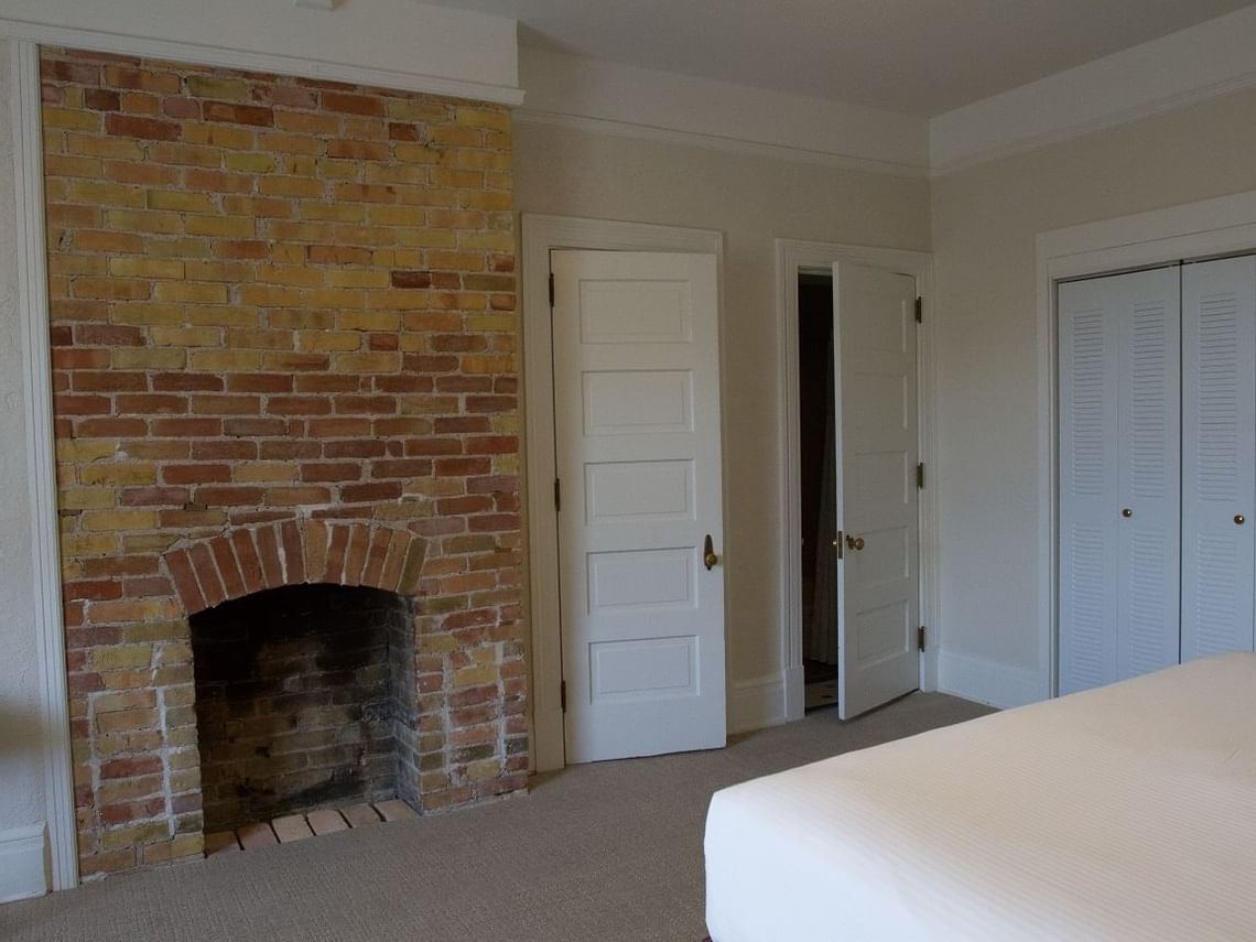 A bedroom with a brick fireplace evokes a sense of naval history, accompanied by two white doors and a closed louvered closet. The light-colored walls frame a bed with crisp white linens in the foreground, embodying a simple, clean design reminiscent of ship quarters.