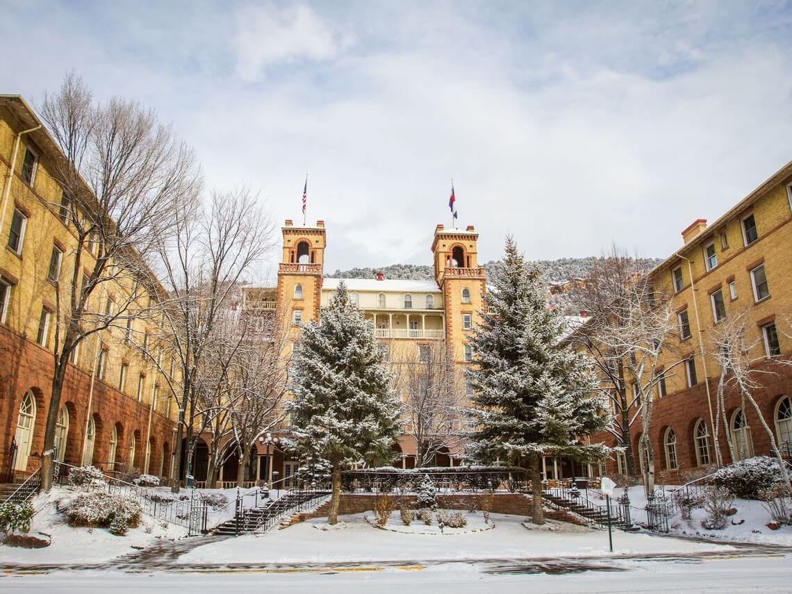 A historic building with two towers stands proudly, framed by snow-covered trees and grounds. The structure’s distinct brickwork and arched windows speak to a reclaimed era. Two flags wave atop the towers against a cloudy sky, adding to its timeless allure.