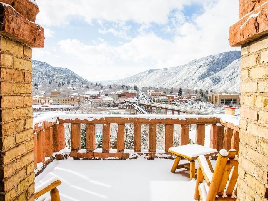 An outdoor snowy balcony with wooden chairs and a table offers a breathtaking view of a town nestled between snow-capped mountains under a partly cloudy sky.