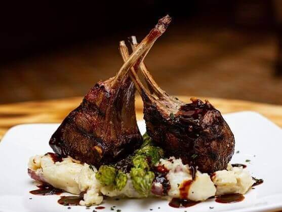 A succulent rack of lamb, expertly crafted by the chef, rests on a bed of creamy mashed potatoes with roasted asparagus, all drizzled with a balsamic glaze on a pristine white plate.