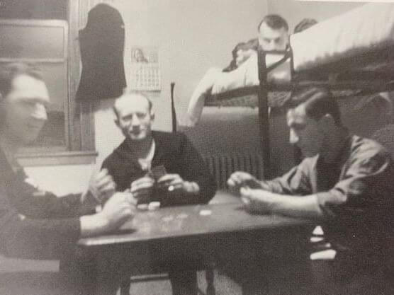 Four men play cards around a small table in a dimly lit room. Two sit facing each other, while the third observes. A fourth man lies on a bunk bed, reminiscent of naval history quarters. A calendar and coat hang on the wall, giving the scene a vintage feel.