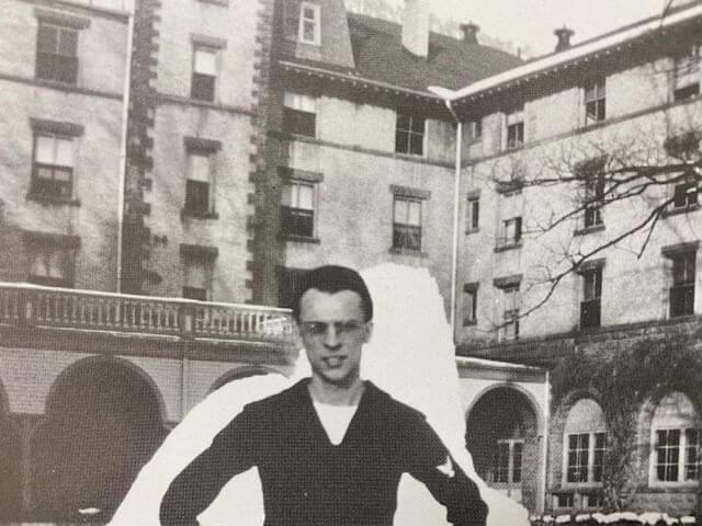 A man in sunglasses and a dark sweater stands before a large, historic building known for its naval history. The black-and-white image captures the structure's detailed architecture, with multiple windows and arched entrances, accompanied by a bare tree on the right.