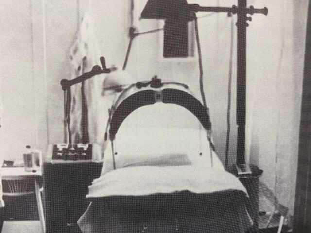 A black and white photo of an old iron lung machine in a medical setting, reminiscent of naval history. The cylindrical apparatus is surrounded by various medical equipment, including lamps and control panels, used historically for respiratory assistance.
