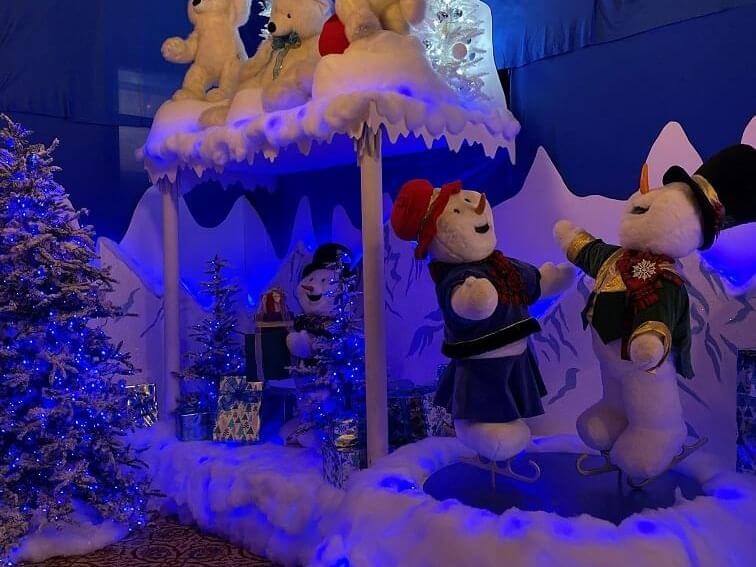 A festive winter scene reimagined with decorated Christmas trees and polar bear plushies on a snowy platform. Two snowman figures, wearing winter clothes, dance joyfully under blue lighting. Gift boxes are scattered around the vibrant display.