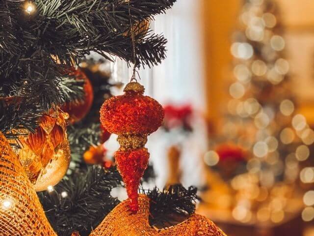 A close-up of a Christmas tree with a red and gold ornament hanging. Warm bokeh lights illuminate the background, creating a cozy and festive atmosphere reminiscent of a winter wonderland.