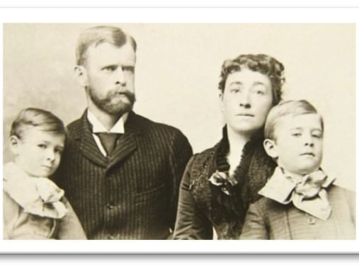 A vintage sepia-toned portrait of a family, steeped in haunted history. Two young boys in matching attire with large bows stand beside a man with a mustache and beard and a woman with curly hair, all clad in formal clothing, gazing toward the camera, their expressions echoing whispers of the past.