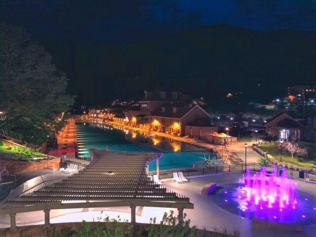 A nighttime scene of a resort pool area offers stunning photo opportunities, illuminated by colorful lights. The foreground features a pergola with lounge chairs, while water fountains glow with purple light. Trees and buildings surround the pool, creating a serene atmosphere.